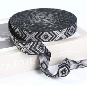 Stacks of multiple colorful custom Jacquard Webbing Tape rolls in different widths (30mm, 40mm) and designs including navy/gold and olive/black on a light grey fabric surface.