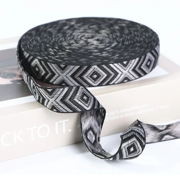 Stacks of multiple colorful custom Jacquard Webbing Tape rolls in different widths (30mm, 40mm) and designs including navy/gold and olive/black on a light grey fabric surface.